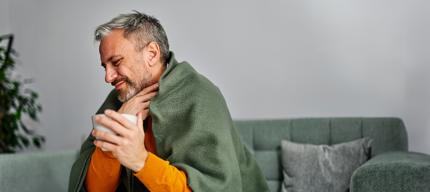Ein Mann mit einer Teetasse in der Hand sitzt in eine Decke eingehüllt auf dem Sofa und greift sich mit schmerzverzerrtem Gesicht an den Hals