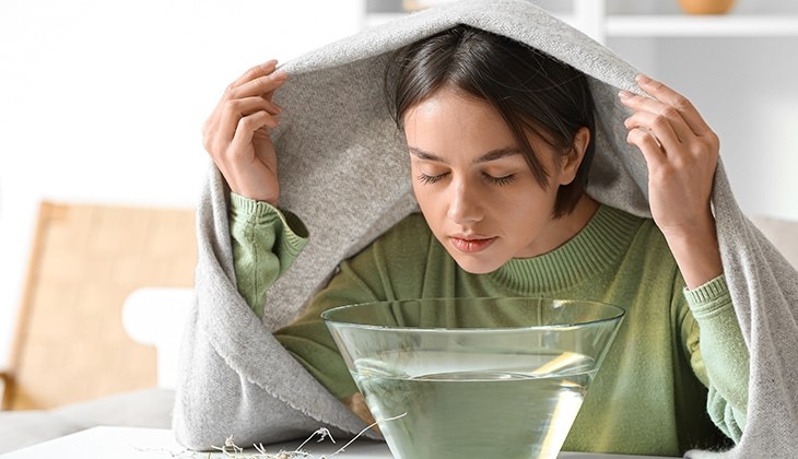 Eine junge Frau lehnt sich zum Inhalieren mit geschlossenen Augen und Tuch über dem Kopf über eine Schüssel mit heißem Wasser und Kamille