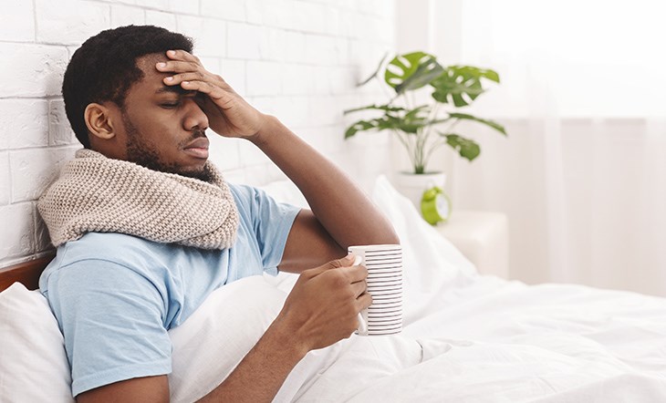 Ein Mann liegt mit einer Tasse Tee im Bett und haelt seine linke Hand an seine Stirn