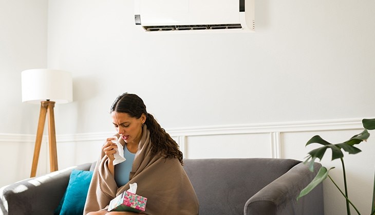 Eine Frau sitzt mit einer Decke umgewickelt und Taschentüchern auf dem Schoß auf der Couch unter einer Klimaanlage und