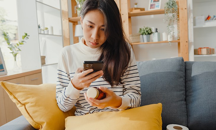 Eine Frau sitzt auf der Couch. In der linken Hand hält sie eine Medikamentenflasche, in der rechten ihr Handy, auf welches sie schaut.