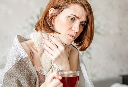 Eine rothaarige Frau mit Schal umgebunden, hält sich den Hals vor Schmerzen und trinkt Tee