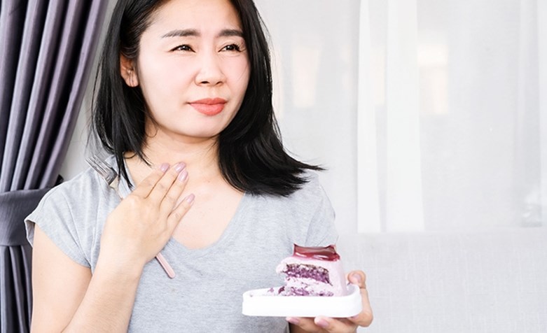 Eine Frau mit dunklem Haar hält in der linken Hand einen Teller mit Kuchen und mit der linken Hand fasst sie sich an in ihren Hals