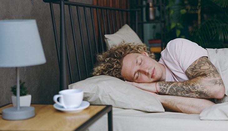 Eine tätowierte Person liegt mit geschlossenen Augen im Bett, auf dem Nachttischschrank ist eine Teetasse
