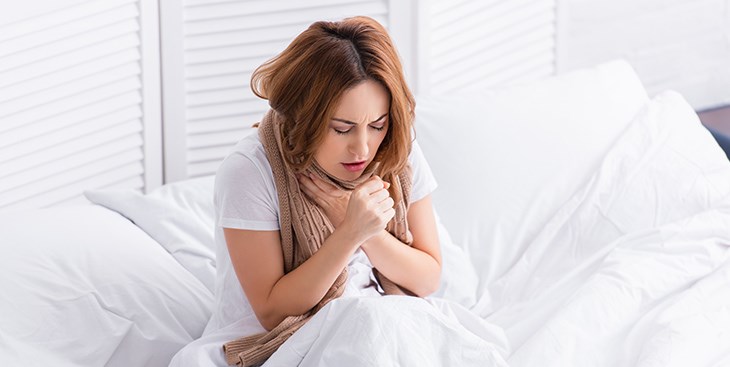 Eine Frau sitzt im Bett und fasst sich an ihren Hals waehrend sie hustet