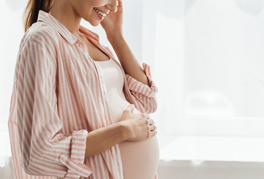 Eine Schwangere Frau hält lächelnd ihre rechte Hand an ihren Bauch