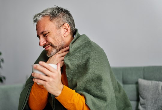 Ein Mann mit einer Teetasse in der Hand sitzt in eine Decke eingehüllt auf dem Sofa und greift sich mit schmerzverzerrtem Gesicht an den Hals