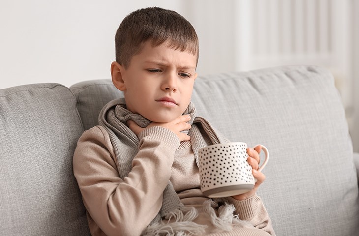 Junge mit Schal sitzt auf einem Sofa, hält eine Tasse in der einen Hand und fasst sich mit der anderen Hand an den Hals
